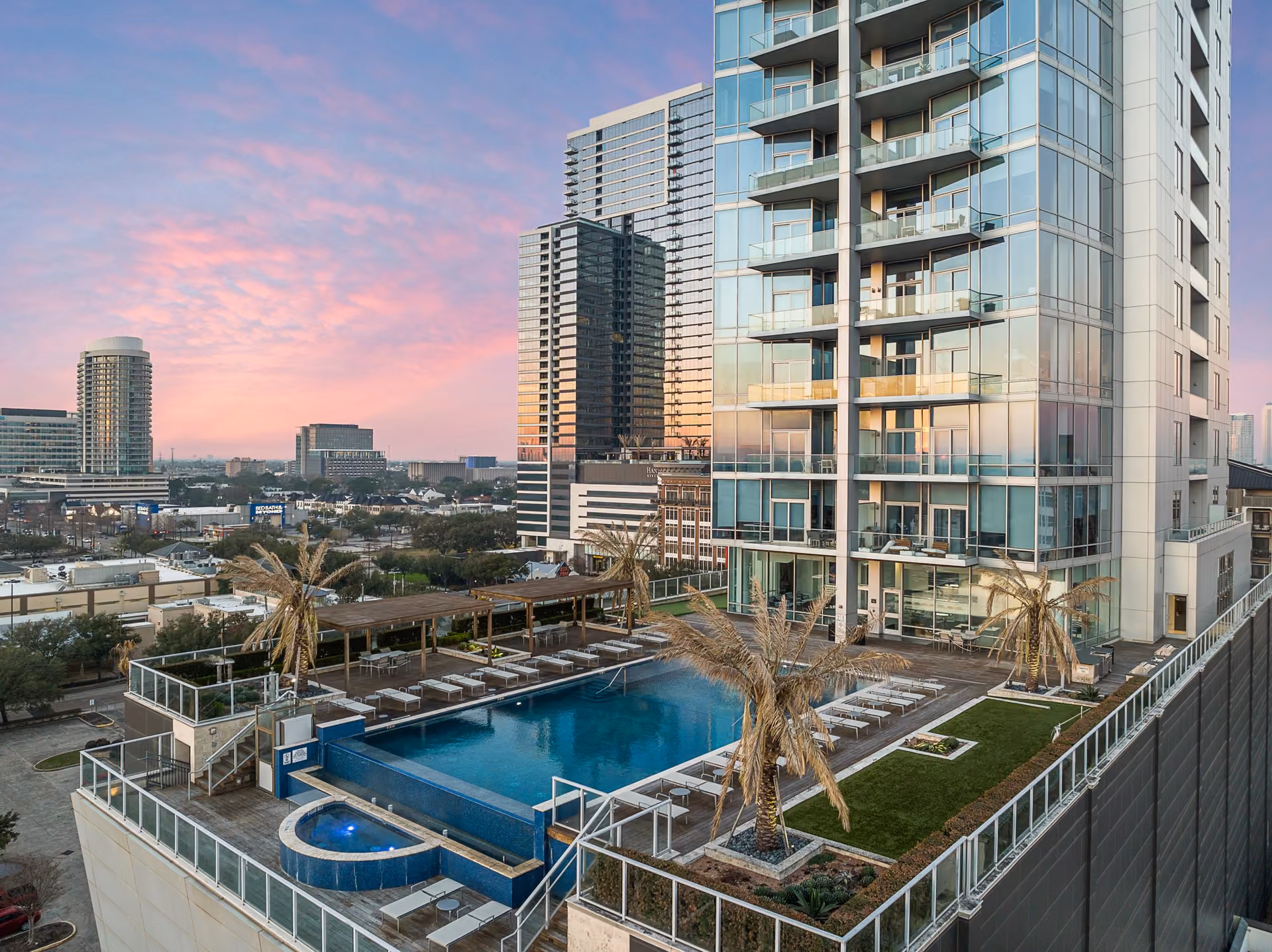 Evening view over the pool terrace at 2727 Kirby: a rectangular blue pool and a circular spa on a raised deck, rows of white loungers, a wooden pergola with dining tables, palm trees and a strip of lawn, the tower's glass balconies rising on the right, and Houston mid-rise and high-rise buildings under a pink and purple sky.